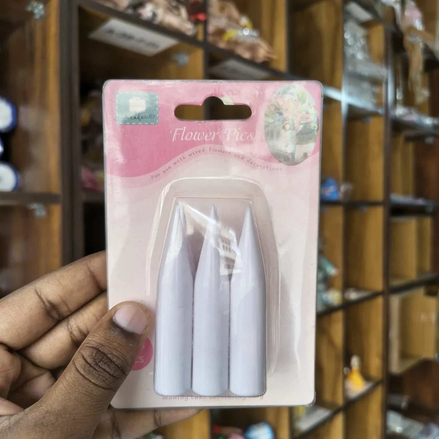 A hand holding a blister pack containing three white plastic flower picks against a blurred wooden shop background.