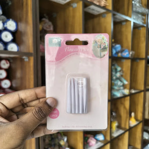 A hand holding a blister pack of white cake decorating picks in a shop setting with wooden shelves in the background.