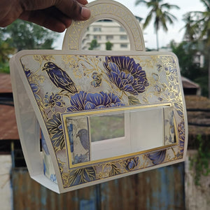 A hand holds a decorative handbag with a floral and bird pattern in blue, gold, and white, set against an outdoor background with palm trees and a building.
