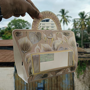 A hand holds a structured handbag-shaped box with a floral print, clear front window, and gold trim, photographed outdoors with buildings and palm trees in the background.