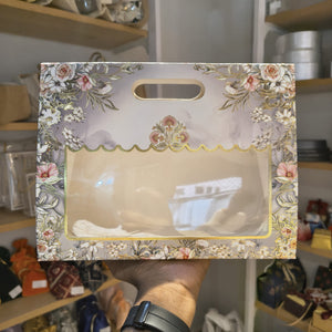 A hand holds a decorative gift box with a floral border and a clear plastic window, showing packaged items inside. The background shows shelves with various items in a retail or storage environment.