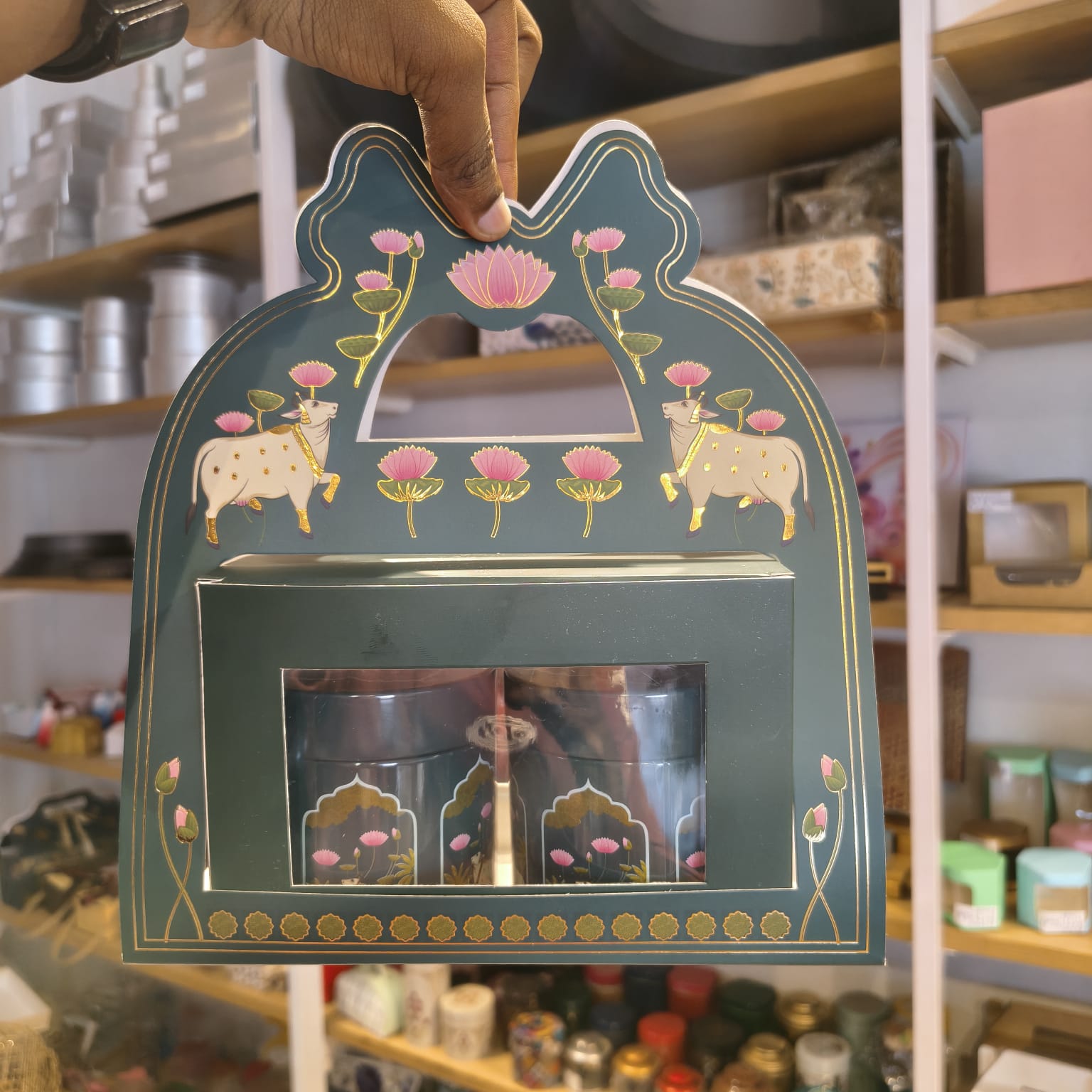 A hand holds a decorative teal gift box with gold and pink illustrations of cows, lotus flowers, and a central window displaying two small glass jars. The background shows a shop interior with shelves and jars.