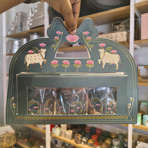 A hand holds a decorative teal gift box with gold and pink hand-painted motifs, including two cows and lotus flowers, with clear plastic bags visible inside.