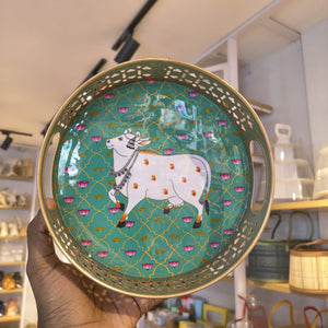 A hand holds a round decorative tray with a white cow illustration on a turquoise patterned background, featuring a gold-toned rim and cutout edges, in a shop setting.