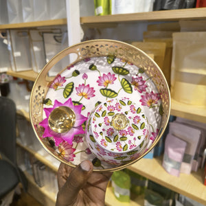 A hand holds a round tray with a matching small lidded container. The tray and container are decorated with a pink and green lotus floral pattern on a white background, with a gold-toned metal rim and a gold-toned lid knob.