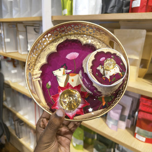 A hand holds a decorative round tray and matching cup set with a magenta floral design, white cow illustration, and gold metallic accents in a retail store setting.