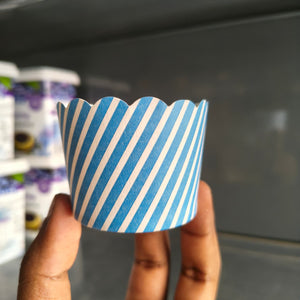A hand holding a paper cup liner with diagonal blue and white stripes, set against a blurred background of what appears to be a refrigerator or cooler shelf.