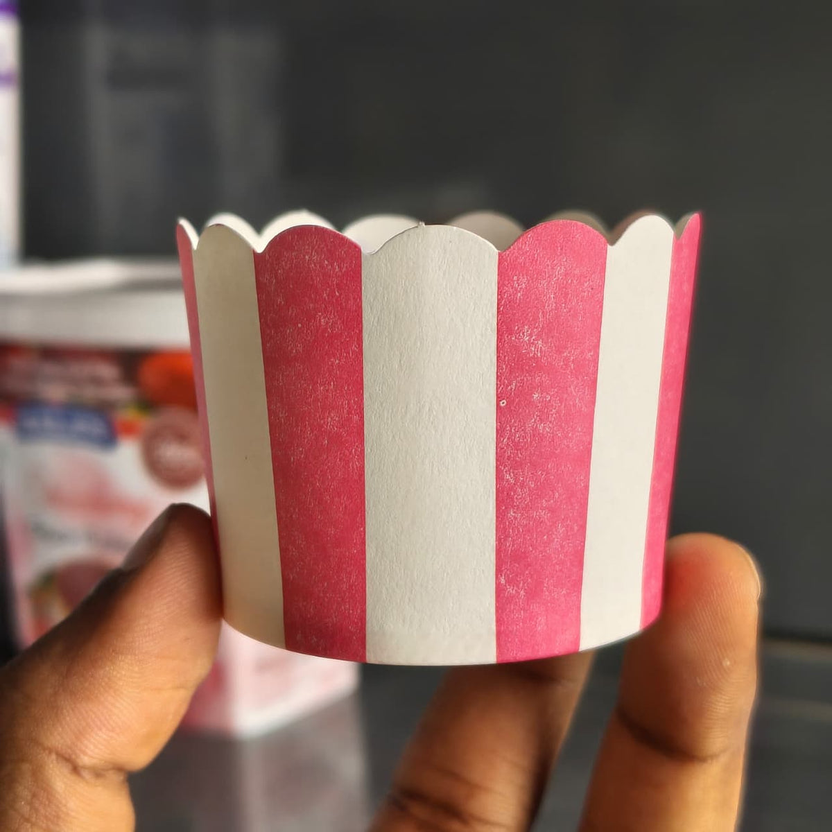 A hand holding a paper cupcake liner with vertical pink and white stripes, set against a blurred background.