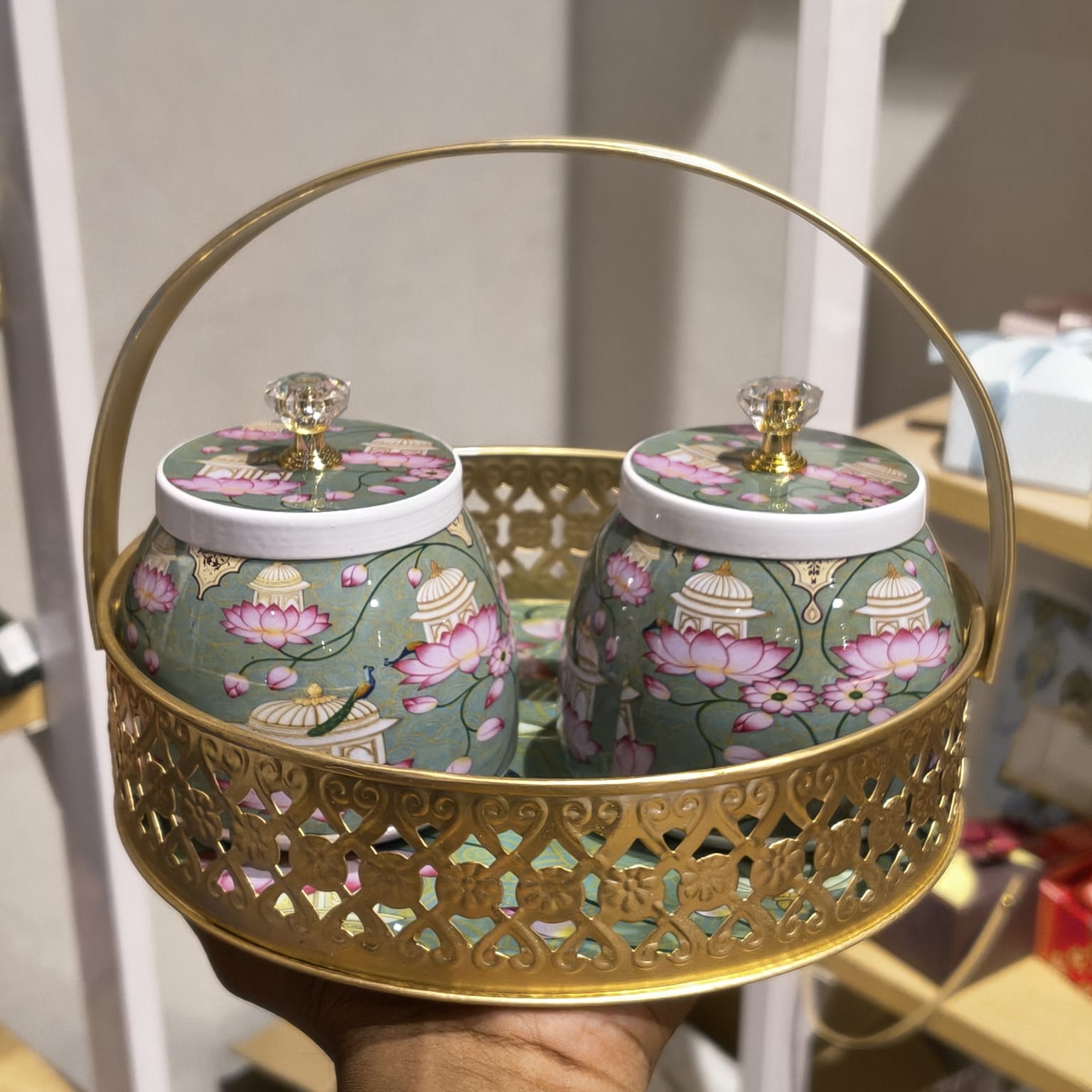 A decorative gold metal carrying basket with two matching ceramic jars featuring pink lotus and pavilion motifs on a sage green background, held in a hand.