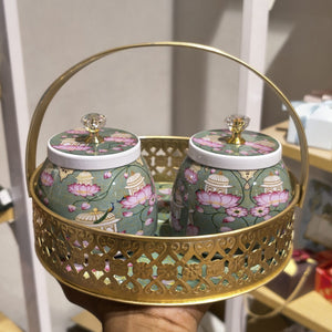 A decorative gold metal carrying basket with two matching ceramic jars featuring pink lotus and pavilion motifs on a sage green background, held in a hand.