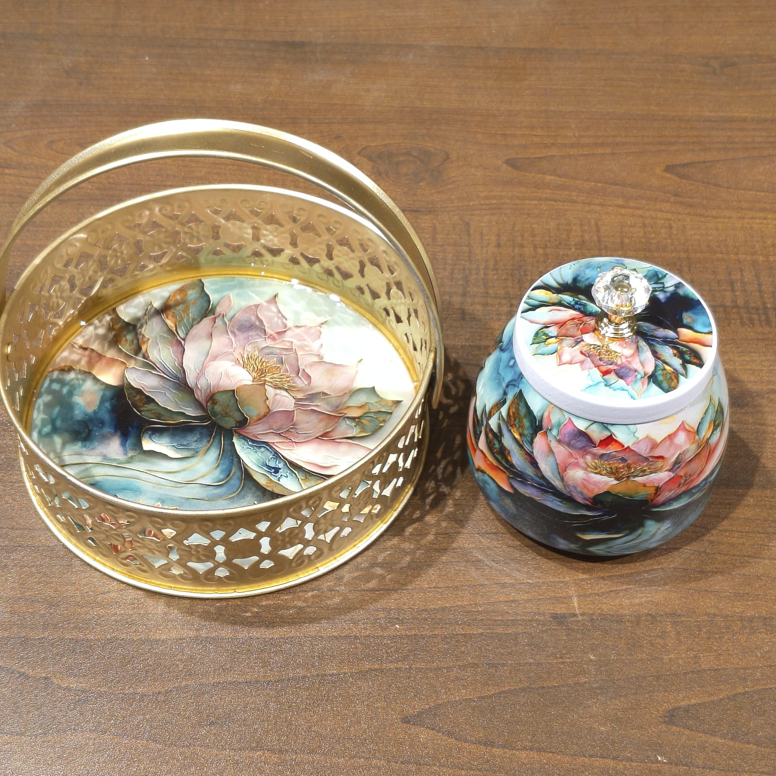 A decorative gold metal basket with a floral hand-painted interior sits next to a matching small lidded jar on a wooden tabletop. Both pieces feature colorful lotus and floral artwork in pink, blue, green, and orange tones.