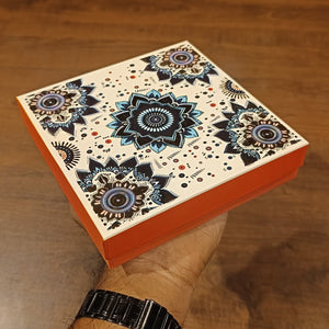 An orange rectangular gift box with a cream lid decorated with intricate black, blue, and brown mandala patterns, held by a hand over a wooden surface.