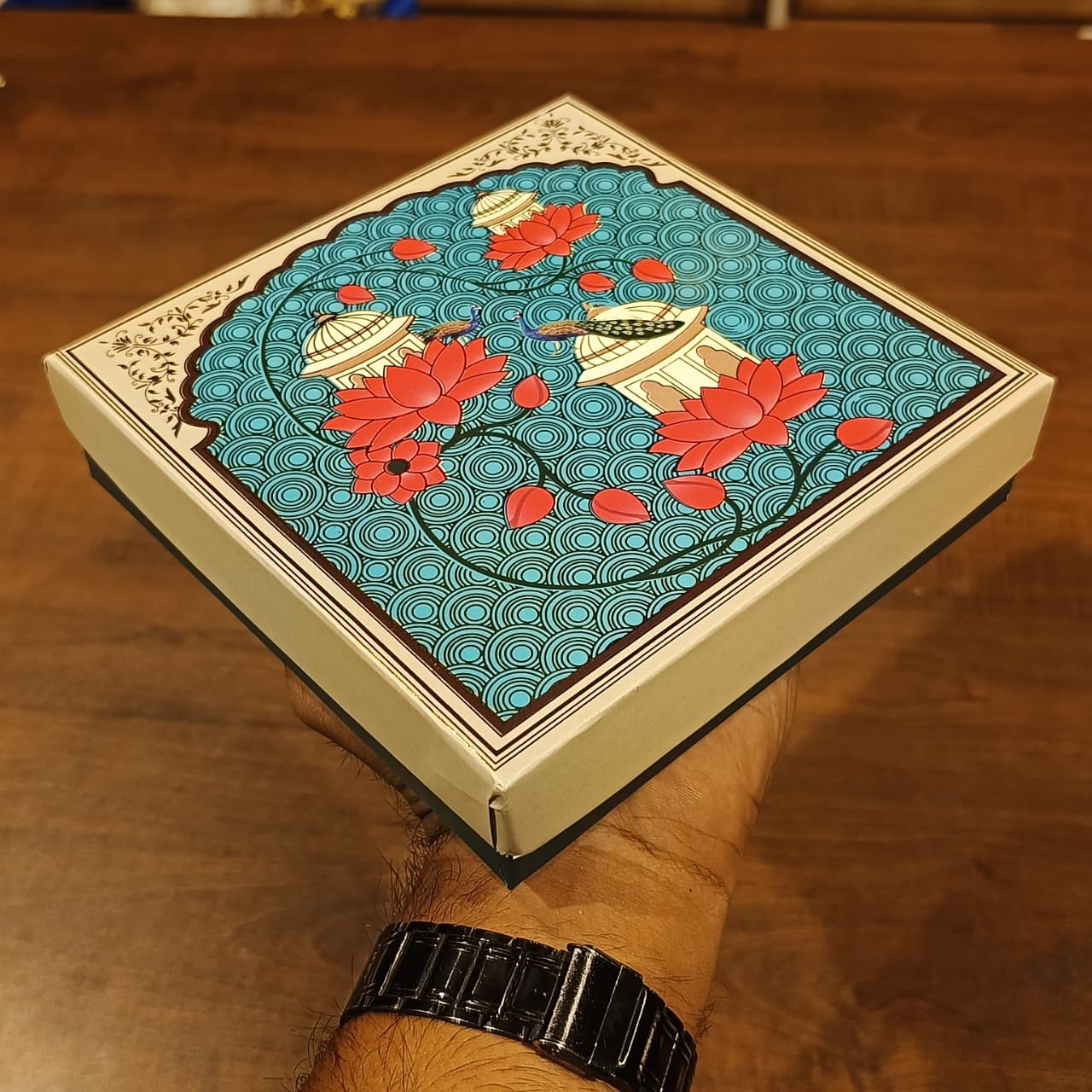 A hand holding a square decorative box with a hand-painted style illustration of red lotus flowers and boats on a turquoise background, set against a wooden table.