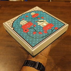 A hand holding a square decorative box with a hand-painted style illustration of red lotus flowers and boats on a turquoise background, set against a wooden table.