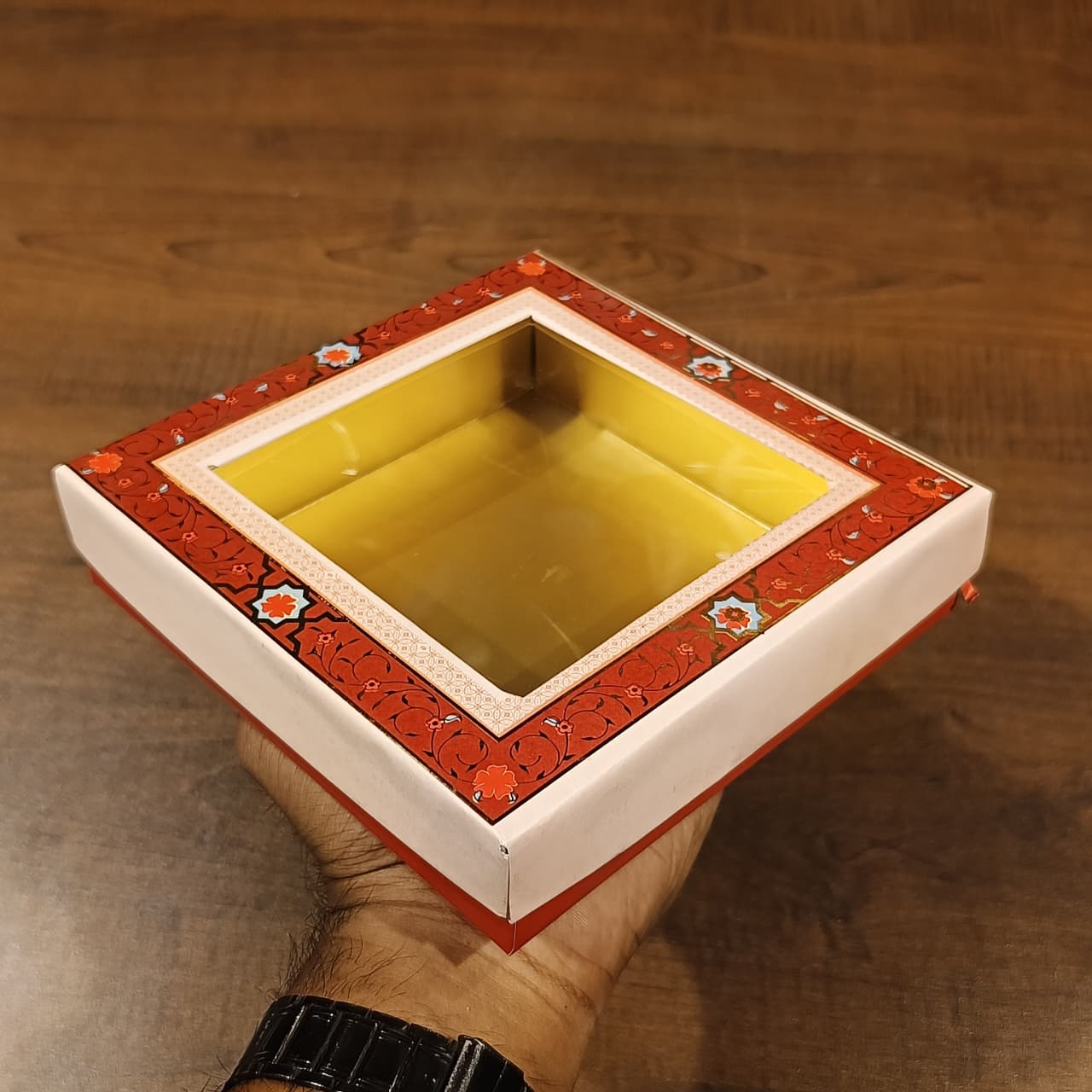 A hand holds a square decorative gift box with a clear top window, featuring a red floral border and a yellow interior, set against a wooden background.