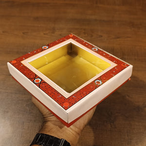 A hand holds a square decorative gift box with a clear top window, featuring a red floral border and a yellow interior, set against a wooden background.