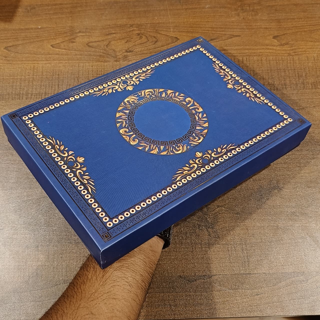 A blue decorative presentation box with ornate gold foil detailing and a central circular motif, held by a person's arm over a wooden tabletop.
