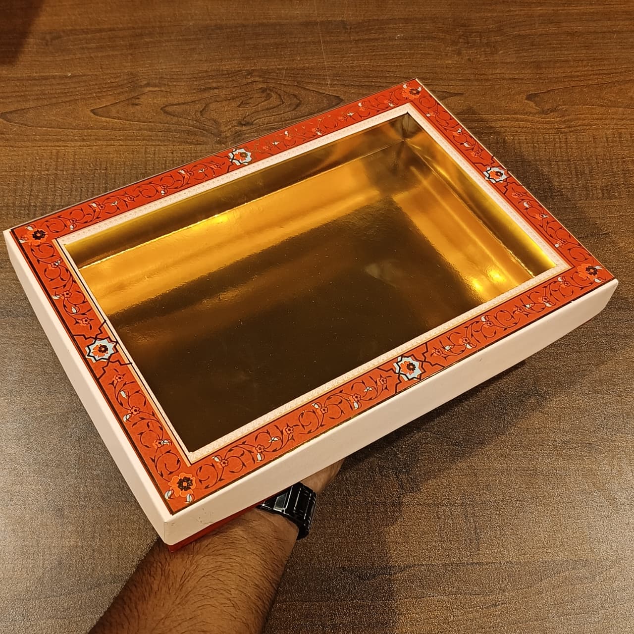 A rectangular decorative tray with a shiny gold interior and an orange-red patterned border, shown resting on a wooden table with a person's arm holding it.