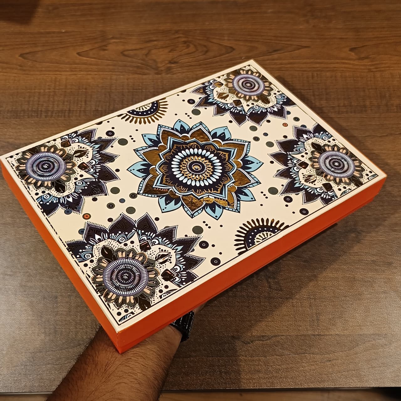 A rectangular decorative box with a cream background and intricate mandala-style floral patterns in navy, black, gold, and blue, held by a person's hand over a dark wood surface.