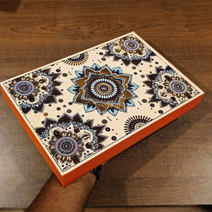 A rectangular decorative box with a cream background and intricate mandala-style floral patterns in navy, black, gold, and blue, held by a person's hand over a dark wood surface.