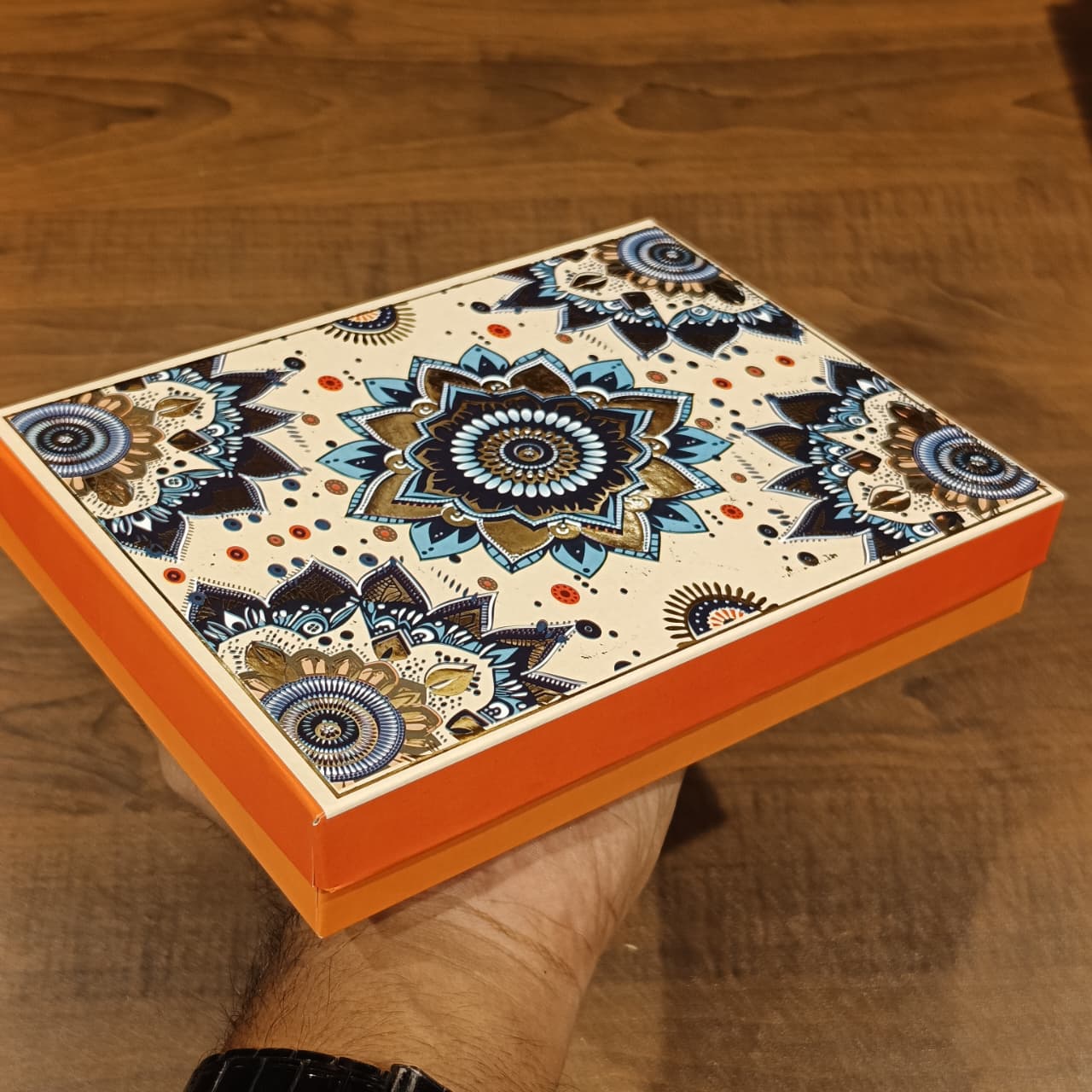 A hand holds a square decorative box with a cream background and an intricate blue, black, and gold mandala pattern on the lid, with bright orange sides, against a wooden tabletop background.