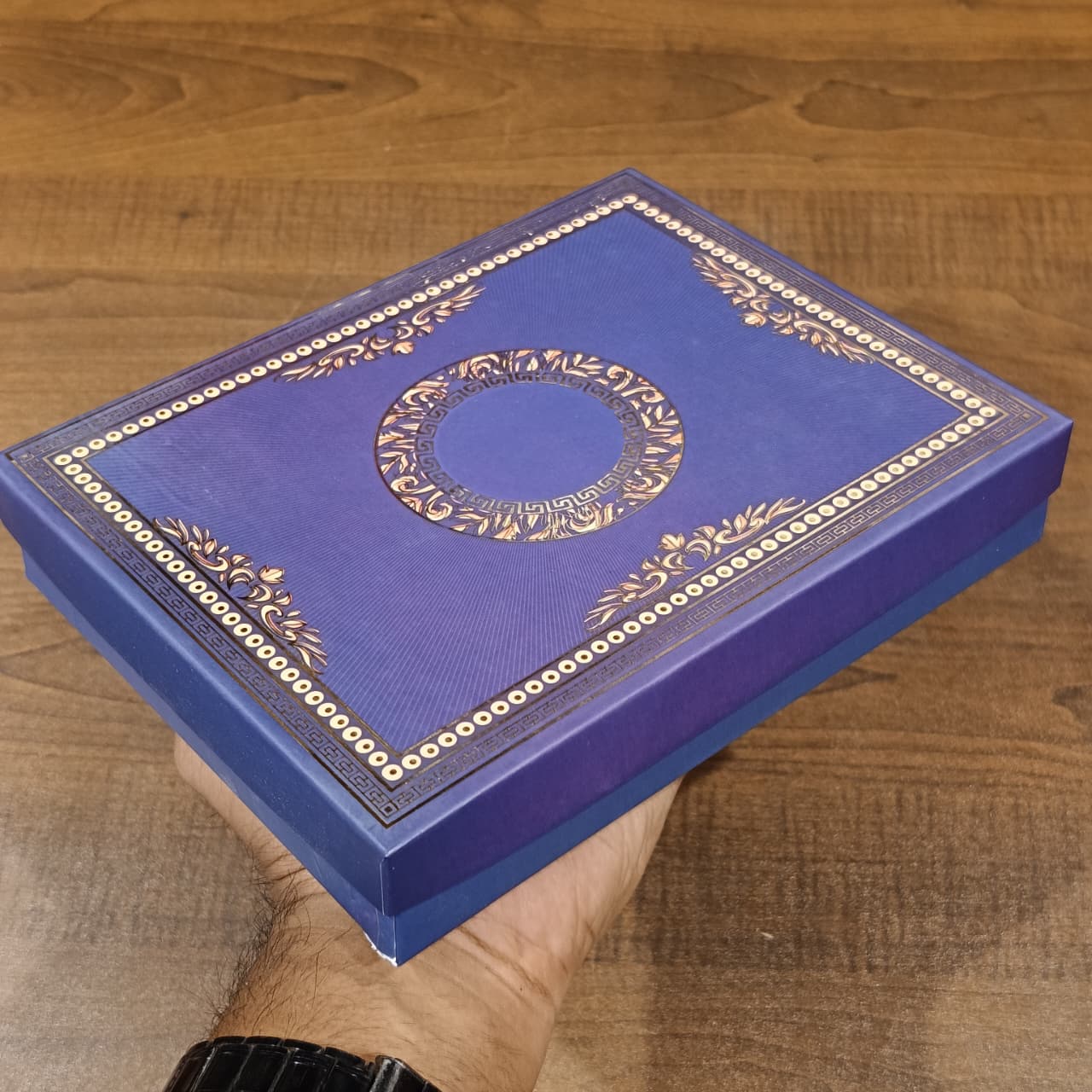 A hand holding a square blue presentation box with ornate gold foil detailing and Arabic calligraphy, set against a wooden table background.