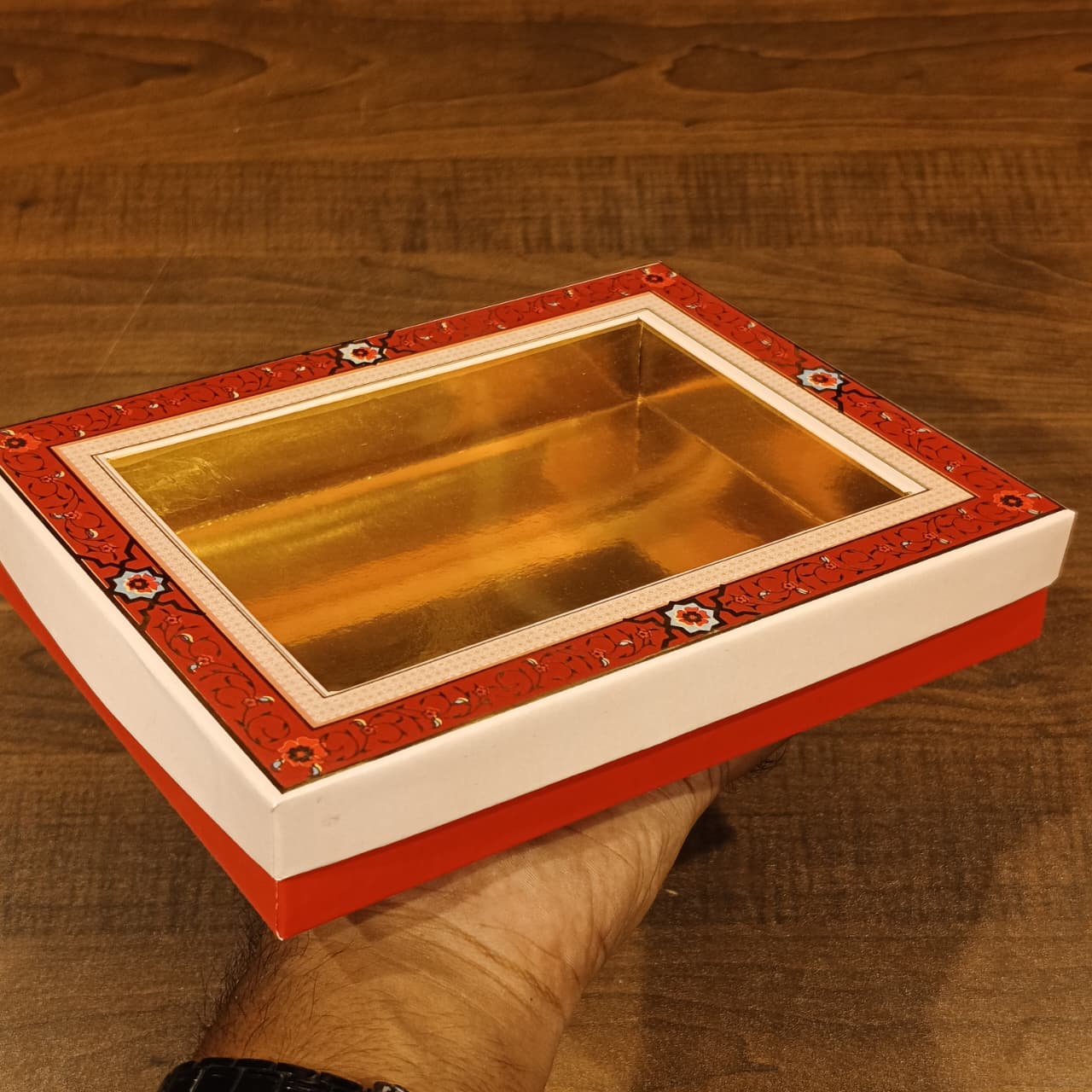 A hand holding a rectangular gift box with a transparent window showing a gold interior, featuring a red patterned border and white sides, set against a wooden background.