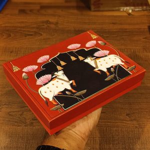 A hand holds a red decorative gift box with a hand-painted style illustration of two white cows under a black arch with pink lotus flowers, set on a wooden table.