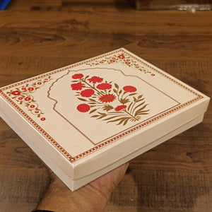 A cream-colored square gift box with red and gold floral illustrations, held by a hand over a rustic wooden table.