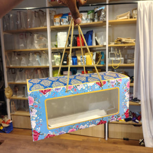 A hand holds a rectangular gift box with a clear front window and floral print. The box has a blue background with pink, red, and white flowers, gold trim, and gold hanging straps, set against a blurred retail store background.
