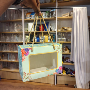 A decorative gift box with a clear front window and gold rope handles, held by a hand in a retail store setting.