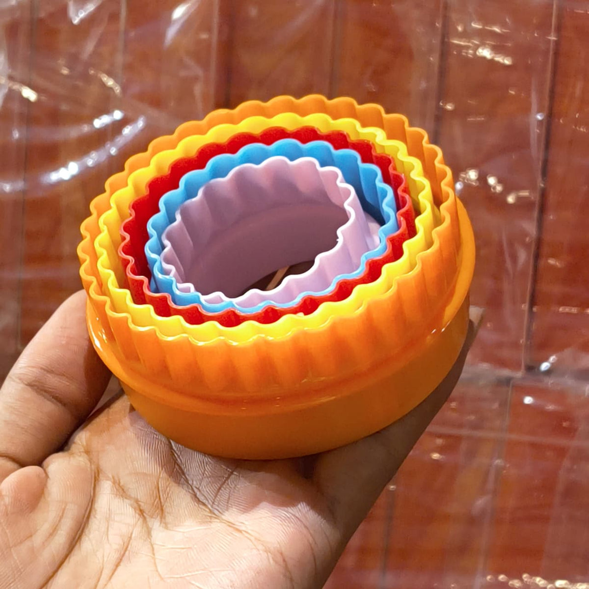 A hand holding a set of nested round plastic ring molds in yellow, orange, red, blue, and pink, photographed against a reflective brown background.