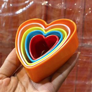 A hand holding a set of nested heart-shaped ceramic dishes in bright colors including orange, white, yellow, blue, and red, with a warm brown background.