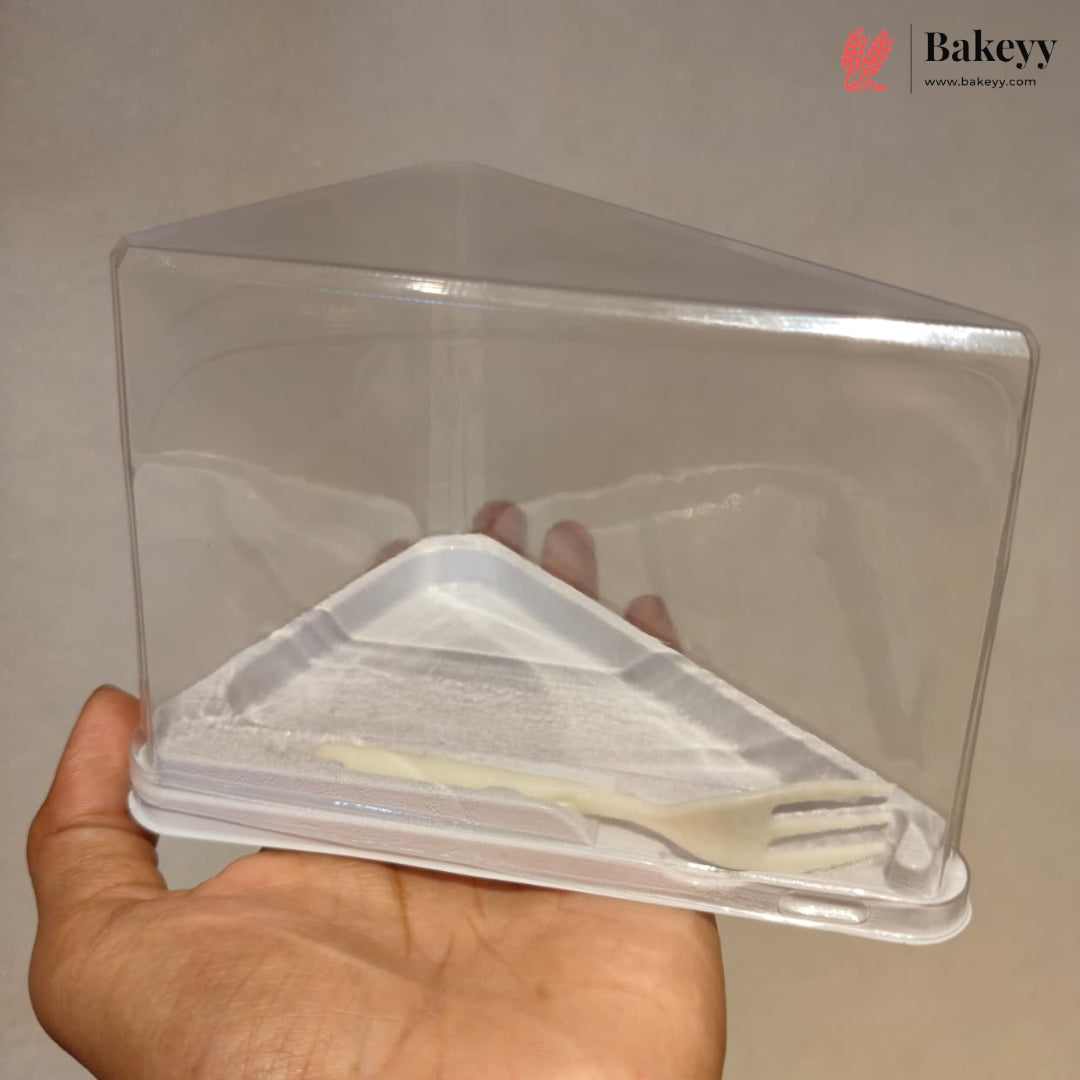 A hand holding a clear plastic triangular cake box with a white plastic insert tray and fork, against a plain beige background. A Bakeyy logo watermark is visible in the top right corner.