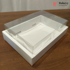 A clear plastic bakery box lid with a white cardboard tray base, shown on a wooden tabletop. A Bakeyy logo watermark is visible in the top right corner.