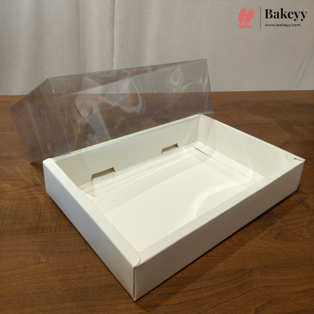 A white rectangular bakery box with a clear plastic lid leaning behind it, placed on a wooden tabletop against a light fabric backdrop. A Bakeyy logo watermark appears in the top right corner.