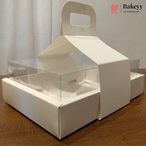 A white cardboard bakery display box with a built-in handle and clear plastic window panels, shown on a wooden tabletop against a light curtain background. A Bakeyy logo watermark is visible in the top right corner.