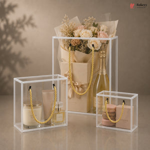 Clear display boxes with gold handles containing a bottle of wine, flowers, and other items on a neutral background.