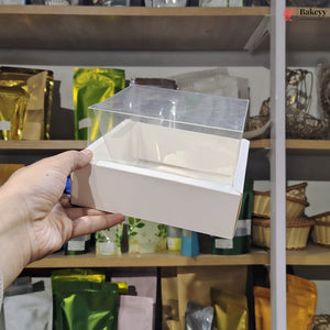 Hand holding a small white box with a clear lid against a blurred store shelf background