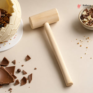 A wooden cake smash hammer with a smooth cylindrical head and long handle, styled on a beige surface with chocolate shards, a partially broken cream cake, and a small bowl of chocolate pieces. Bakeyy logo appears in the top right corner.