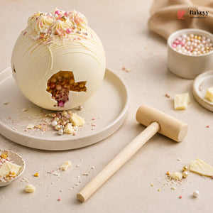 A beautifully styled product photo of a white chocolate egg with a decorative cutout revealing a colorful berry filling, shown on a beige surface with a wooden mallet, white chocolate pieces, and bowls of sprinkles. The Bakeery logo is visible in the top right corner.