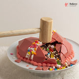 A pink chocolate cake on a white plate with a wooden mallet resting on top, smashing the cake to reveal colorful candy and chocolate pieces inside. The Bakeyy logo is visible in the top right corner.