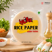 A 400g package of Yoka Rice Paper (Bânh Trâng) displayed on a wooden board with fresh spring rolls, vegetables, and a pepper mill in a kitchen setting.