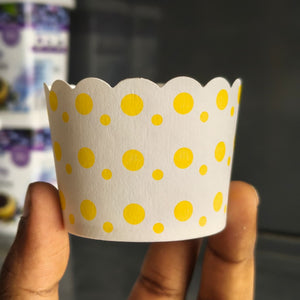 A hand holding a white paper cupcake liner decorated with yellow polka dots, with a blurred background.