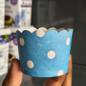 A hand holding a blue paper cupcake liner with white polka dots, with a blurred background showing shelves and products.