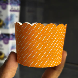 An orange paper cupcake liner with a white diagonal polka dot pattern, held by a hand against a dark, blurred background.