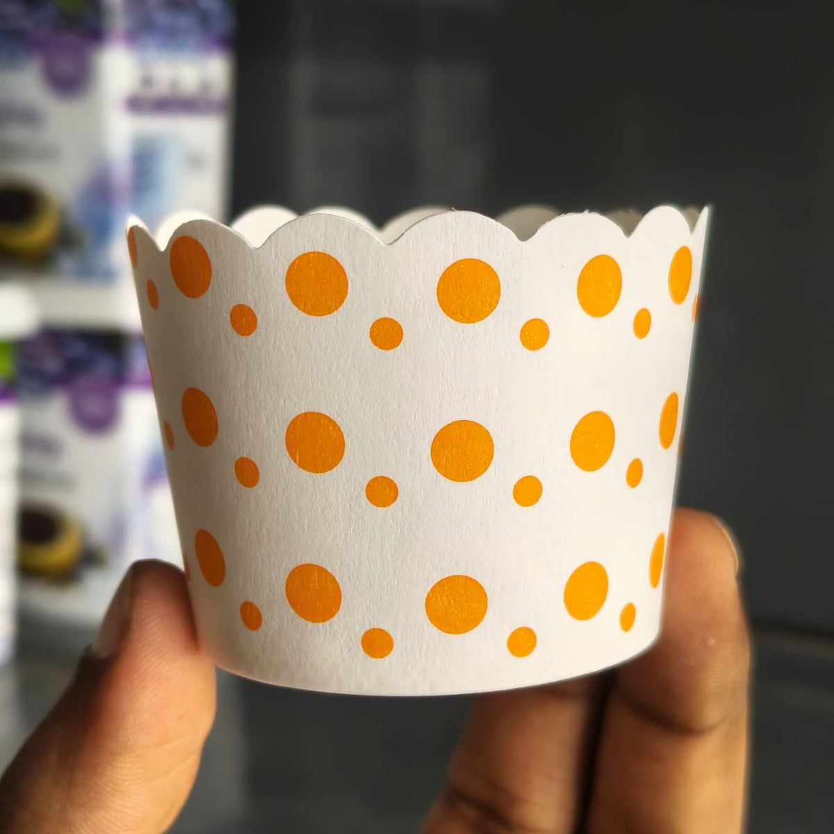 A hand holding a white scalloped paper cup liner decorated with orange polka dots, with a blurred background showing shelves of packaged goods.