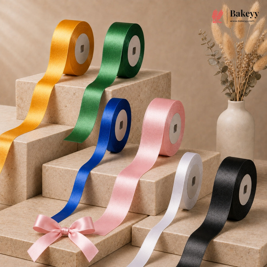 Six rolls of satin ribbon in yellow, green, blue, pink, white, and black, arranged on beige display blocks with a decorative vase in the background. The pink ribbon is styled with a bow.
