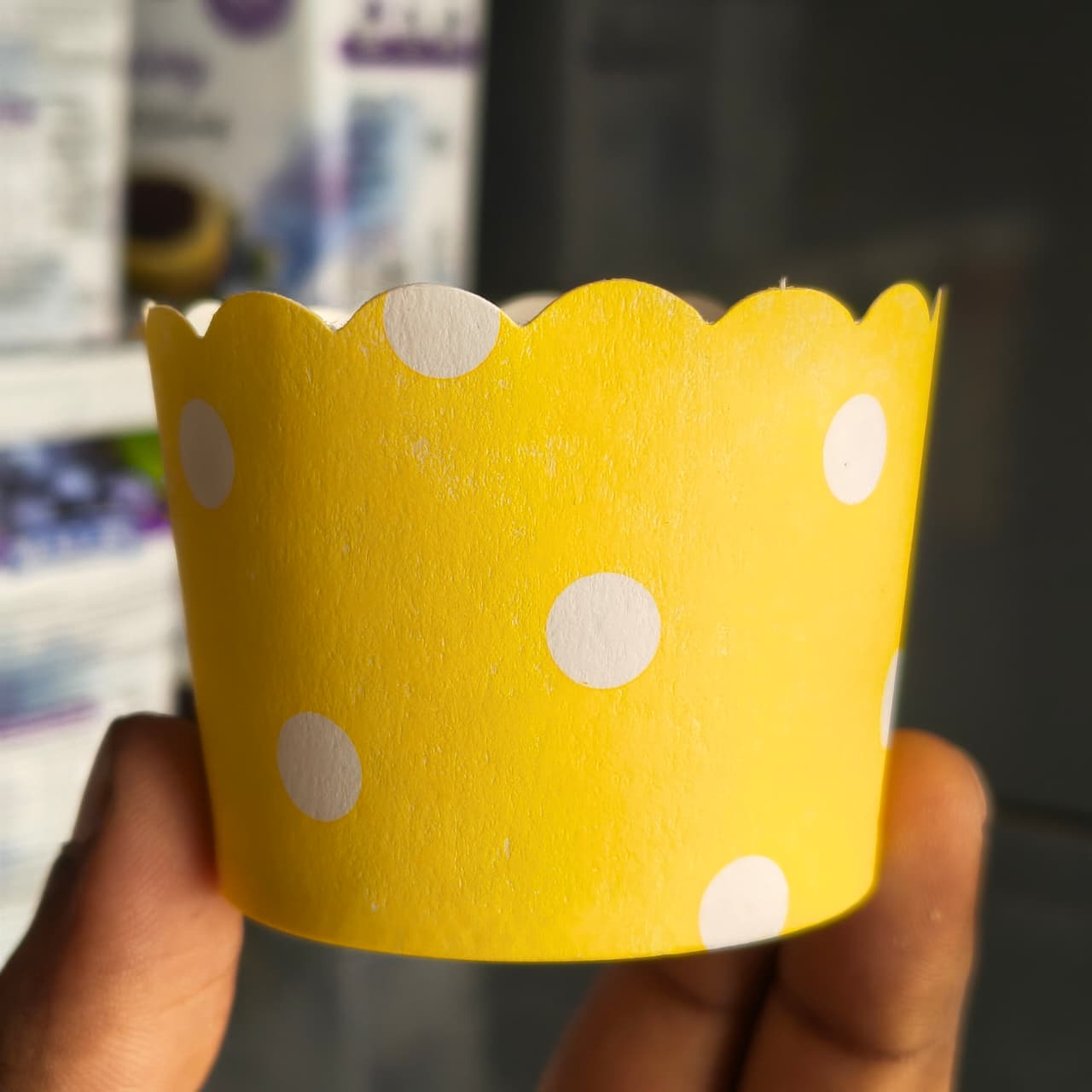 A hand holding a yellow paper cupcake liner with white polka dots, set against a blurred background.