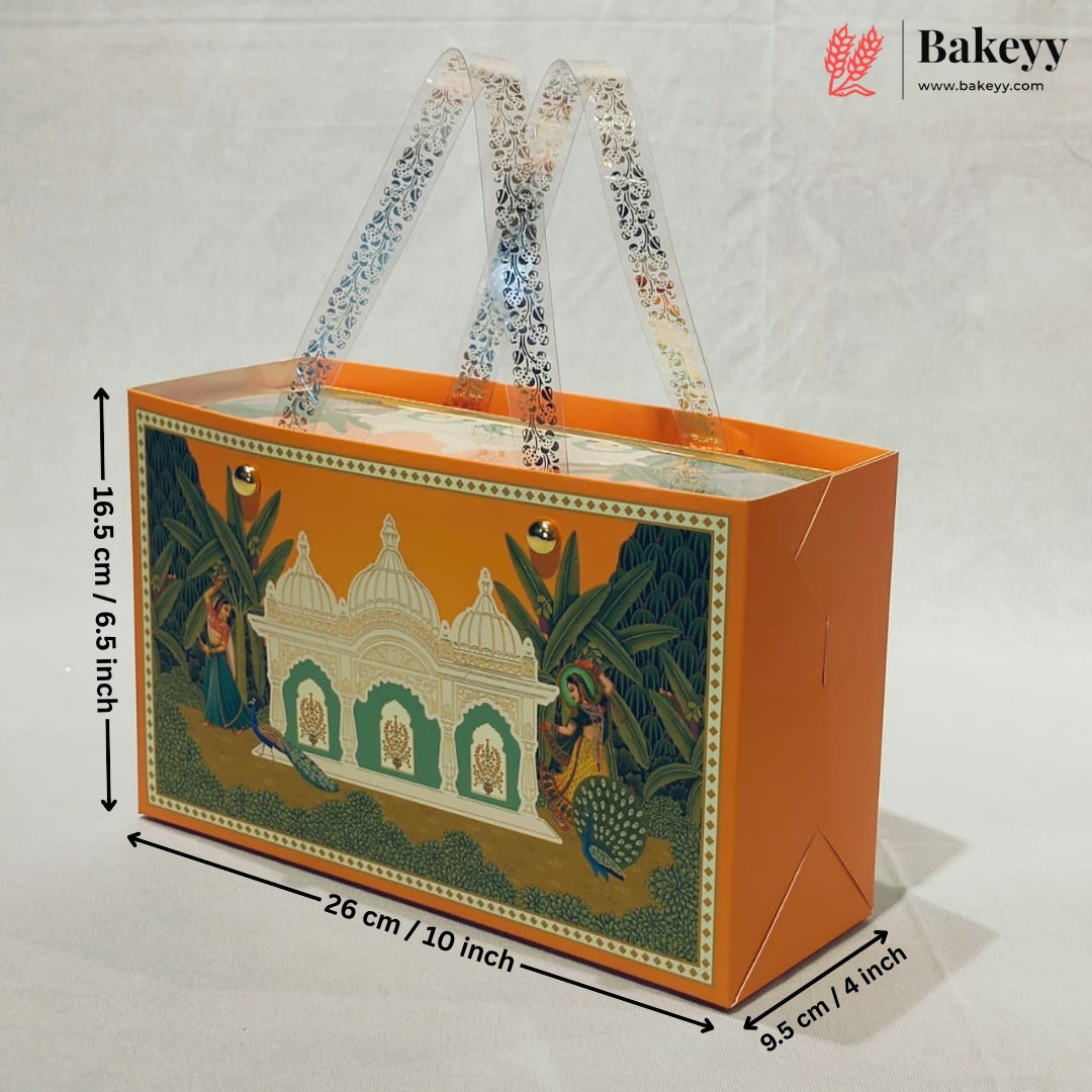 An orange rectangular gift box with clear handles and a printed traditional Indian architectural illustration on the front, shown with dimension arrows and labels indicating 26 cm length, 16.5 cm height, and 9.